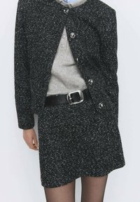 Jachetă din amestec de lână alb-negru cu guler rotund, închidere cu nasturi, asortată cu un pulover gri deschis și o centură neagră.