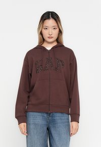 Brown zip-up hoodie featuring a black leopard print "GAP" logo, long sleeves, and a drawstring hood. Soft material with a relaxed fit.