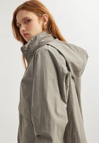 Ljust grå huva jacka med en texturerad finish, med dragkedjestängning och en avslappnad silhuett. Inkluderar synlig sömnad och hårdvaruutstick.