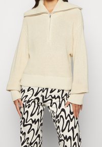 Maglione lavorato a maglia color crema con colletto alto e chiusura a zip, caratterizzato da polsini e orlo a costine. Abbinato a pantaloni neri e bianchi con motivo ondulato.