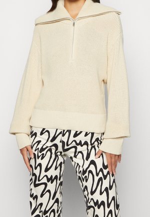 Cremfarbener Strickpullover mit hohem Kragen und Reißverschluss, ausgestattet mit gerippten Bündchen und Saum. Kombiniert mit schwarz-weißen Hosen mit wellenförmigem Muster.