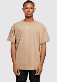 Kortærmet, sandbeige bomulds-T-shirt med rund hals og hvid tekst "MERCHC+DE" på brystet. God pasform og ensfarvet.