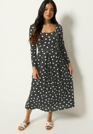 Kobieta w czarnej sukience z długim rękawem w białe grochy i białe płaskie sandały, stojąca na jednolitym jasnym tle.