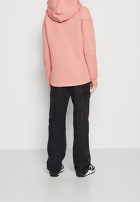 Übergroßer pinkfarbener Hoodie mit Kapuze, kombiniert mit dunklen Cargohosen mit seitlichen Taschen, vor einem schlichten hellen Hintergrund.