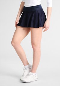 Falda deportiva azul marino con diseño acampanado, hecha de tela transpirable. Cuenta con una cinturilla ancha y una longitud extendida. Se lleva con zapatillas blancas.