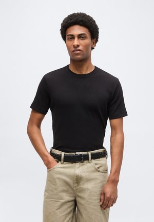 Mann mit Afro-Frisur, der ein schwarzes kurzärmeliges Shirt und beigen Hosen mit einem geflochtenen schwarzen Gürtel trägt, steht vor einem einfarbigen hellen Hintergrund.