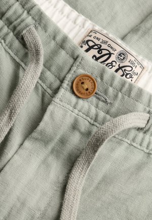 Jasnozielone spodnie z tkaniny z drewnianym guzikiem i beżowym sznurkiem, pokazujące markową metkę wewnątrz pasa.