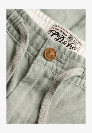 Jasnozielone spodnie z tkaniny z drewnianym guzikiem i beżowym sznurkiem, pokazujące markową metkę wewnątrz pasa.