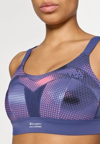 Sujetador deportivo púrpura con patrones de lunares en rosa y negro, con tirantes ajustables y un logo en la parte inferior. Diseñado para la absorción de impactos.