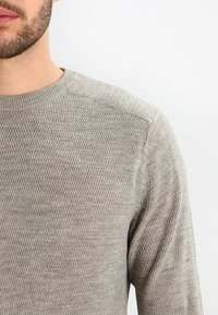 Harmaa neulepaita, jossa on teksturoitu kuvio, pyöreä pääntie ja pitkät hihat. Materiaali on pehmeä ja hieman ribbineulottu.