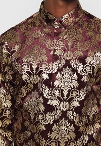 Vínově červená sametová košile s propracovanými zlatými damaskovými vzory, špičatým límečkem a jedním ozdobným knoflíkem u krku.