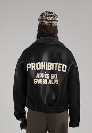 Personne vue de dos porte un bonnet tricoté à motif et une veste en cuir noire portant l’inscription « PROHIBITED APRÈS SKI SWISS ALPS. »