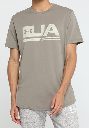 Man draagt een grijs Under Armour T-shirt met een groot "UA" logo en bijpassende grijze joggingbroek met wit Under Armour-logo.