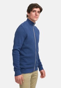 Blå strikket jakke med glidelås og høy krage, som har et teksturert mønster og ribbede mansjetter og kant. Modellen har den på sammen med khakifargede bukser.