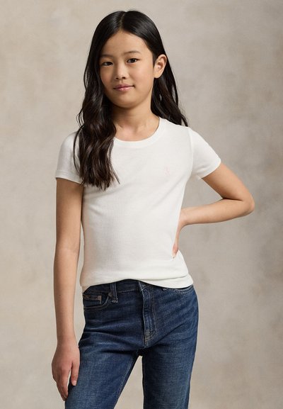 Jeune fille aux longs cheveux foncés, portant un t-shirt blanc à manches courtes et un jean bleu foncé, debout avec une main sur la hanche.