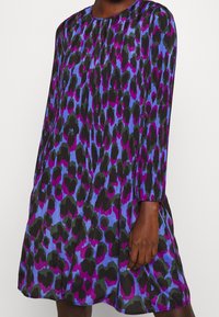 Ein Kleid mit einem schwarz, lila und blauen Leopardenmuster, locker sitzend, langen Ärmeln und rundem Ausschnitt. Strukturierter Stoff und knielang.