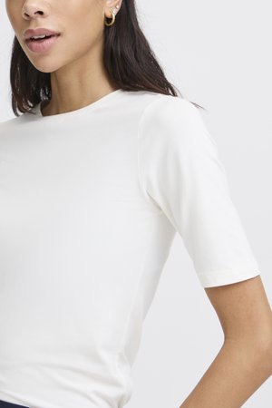 Frau mit einfachem, weißem Kurzarmshirt und goldenen Creolen, mit langen dunklen Haaren vor hellem Hintergrund sichtbar.