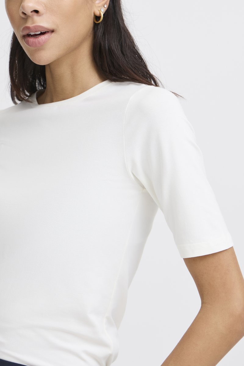 Femme portant un haut blanc uni à manches courtes et une boucle d'oreille créole dorée, avec de longs cheveux foncés visibles sur un fond clair.