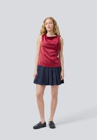 Femme aux longs cheveux blonds ondulés portant un top rouge satiné sans manches, une jupe plissée bleu marine et des mocassins noirs, se tenant sur un fond gris clair.