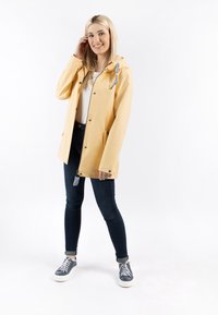 Sárga vízálló dzseki kapucnival, elöl patentos záródással és zsebekkel, sötét farmerral és szürke sportcipővel, fehér talppal kombinálva.