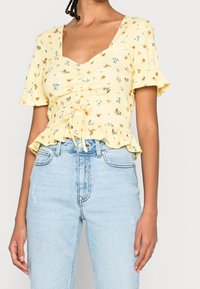 Donna che indossa una blusa floreale gialla a maniche corte con orlo arricciato, abbinata a jeans azzurri a vita alta.