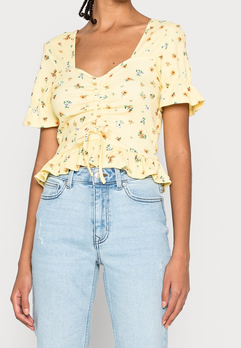 Donna che indossa una blusa floreale gialla a maniche corte con orlo arricciato, abbinata a jeans azzurri a vita alta.