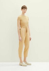 Senfgelbes kurzärmeliges Oberteil kombiniert mit hoch taillierten, locker sitzenden beigen Hosen und cremefarbenen Plateauschuhen, vor einem hellen Hintergrund.