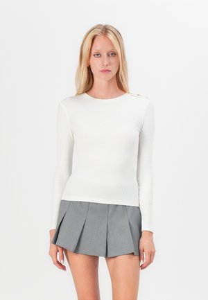 Hvit langermet ribbet topp med rund hals og dekorative knapper på den ene skulderen, kombinert med et grått plissé mini-skjørt.