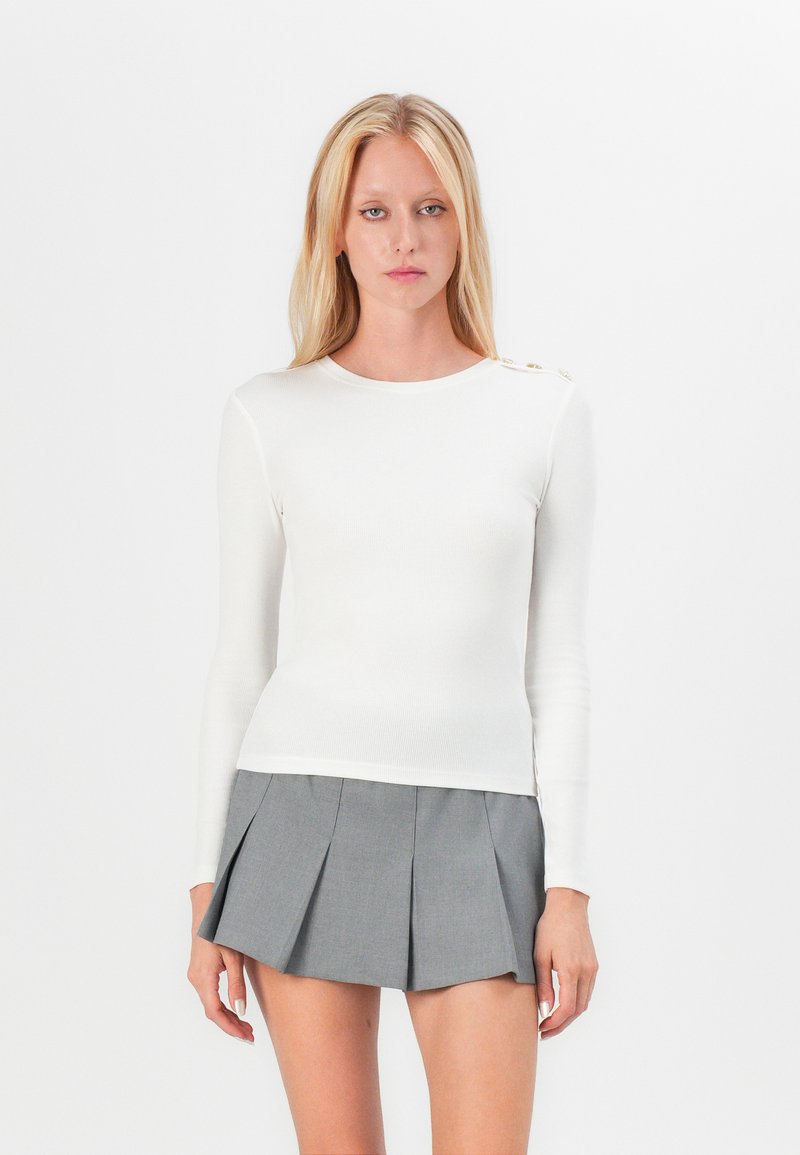 White long-sleeve ribbed top with crew neck and decorative buttons on one shoulder, paired with a gray pleated mini skirt.