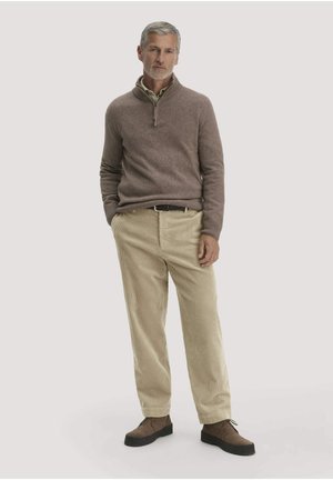 Mann steht, trägt braunen Pullover mit Viertelreißverschluss, beige Cordhosen und braune Schuhe vor hellem einfarbigem Hintergrund.