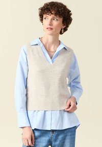 Beige, gestrickte Weste mit V-Ausschnitt über einem hellblauen Hemd mit Kragen, ausgestattet mit einem gerippten Saum, getragen mit lockeren blauen Jeans.
