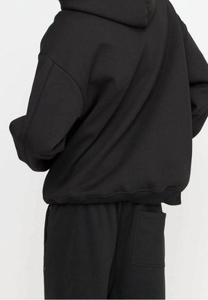 Personne portant un sweat à capuche noir ample et un pantalon noir assorti avec une poche arrière, se tenant devant un fond uni.