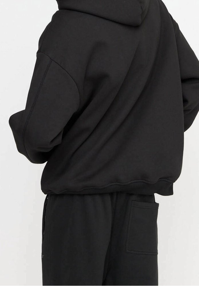 Personne portant un sweat à capuche noir ample et un pantalon noir assorti avec une poche arrière, se tenant devant un fond uni.
