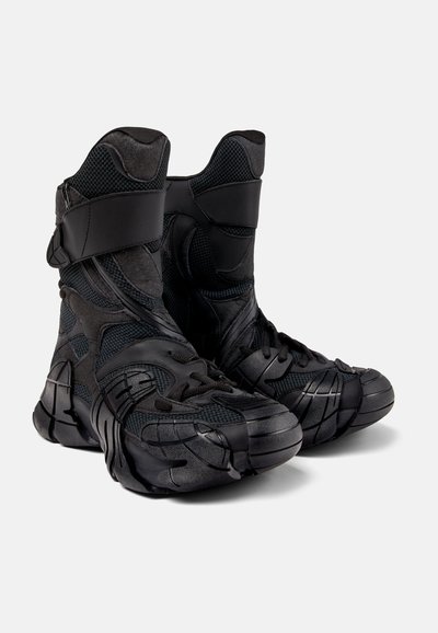 Svarta högskurna stövlar gjorda av texturerat nät och syntetiskt material. Har kurviga detaljer, justerbart rem och gummisulor med framträdande mönster.