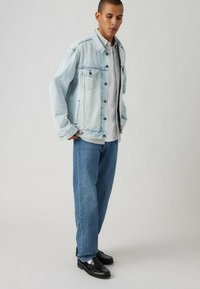 Giacca di jeans azzurro chiaro con due tasche sul petto, abbinata a una camicia bianca e jeans blu. Mocassini neri completano il look.