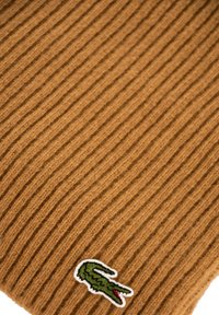 Tessuto a maglia marrone a coste con un piccolo logo coccodrillo verde ricamato vicino al bordo inferiore.