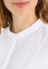 Blouse blanche à encolure arrondie, présentant des plis verticaux et de subtils petits pois texturés. Détail de bouton au col. Tissu léger et aérien.