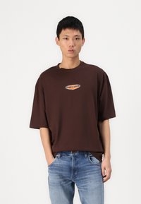 Camiseta oversized marrón con escote redondo y un logo central. Combinada con unos vaqueros de denim azul claro, de corte clásico y textura desgastada.