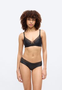 Γυναίκα με σγουρά μαλλιά, που φοράει μαύρο σουτιέν με δαντέλα και ταιριαστό εσώρουχο, στέκεται μπροστά σε απλό ανοιχτογκρι φόντο.