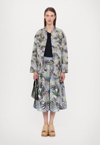 Blümchenjacke in hellen Farben, kombiniert mit einem passenden Rock. Eine schwarze Handtasche und Schuhe mit Holz-Plateausole runden das Outfit ab.