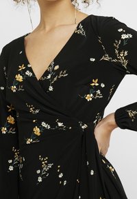 Svart blommig klänning med knytdesign, prydd med gula och vita blommor, lång ärm och draperad sidodetalj. Mjuk tygtextur.