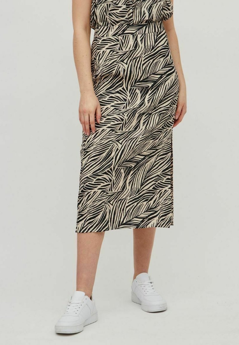 Femme portant une jupe midi noire et beige à imprimé zèbre avec des baskets blanches, debout devant un fond clair uni.