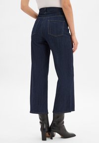 Jeans z wysokim stanem i szerokimi nogawkami w ciemnoniebieskim kolorze z dwoma tylnymi kieszeniami i kontrastującym szwem, zestawione z czarnymi botkami na obcasie.