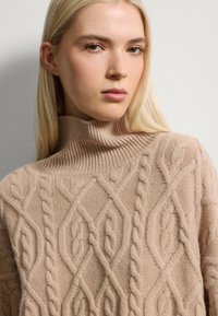 Suéter beige de punto cableado con un alto cuello acanalado, que presenta patrones intrincados y una textura suave. La vista en primer plano destaca la artesanía.