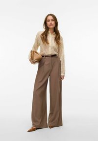 Kreemi värvi pitspluus nööpidega, kombineeritud kõrge vöökohaga, laia säärega pruunide pükstega ja beeži käekotiga, neutraalse taustaga.