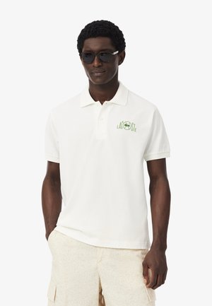 Homme portant des lunettes de soleil noires, un polo blanc Lacoste avec un logo vert, et un short cargo de couleur claire, debout les mains dans les poches.