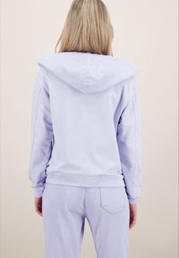 Světlofialová mikina s velkou kapucí, dlouhými rukávy a volným střihem. Měkká látka s jemnými švy; v kombinaci s odpovídajícími kalhotami.
