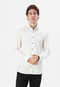 Chemise blanche à boutons noirs, à manches longues et col classique. Présente un détail contrastant à l'intérieur du col et une texture lisse.