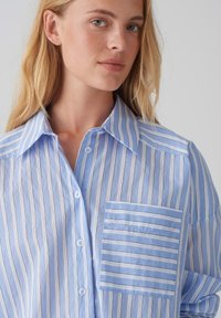 Camicia a righe azzurro chiaro e bianca in cotone, con bottone anteriore, grande taschino sul petto e design con colletto largo.