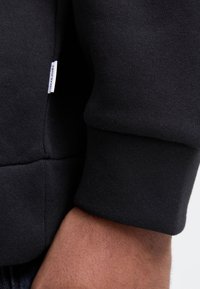 Sort sweatshirtmateriale med ribbede manchetter, der viser et nærbillede af kanten og mærkedetaljen. Glat tekstur, enkelt design.